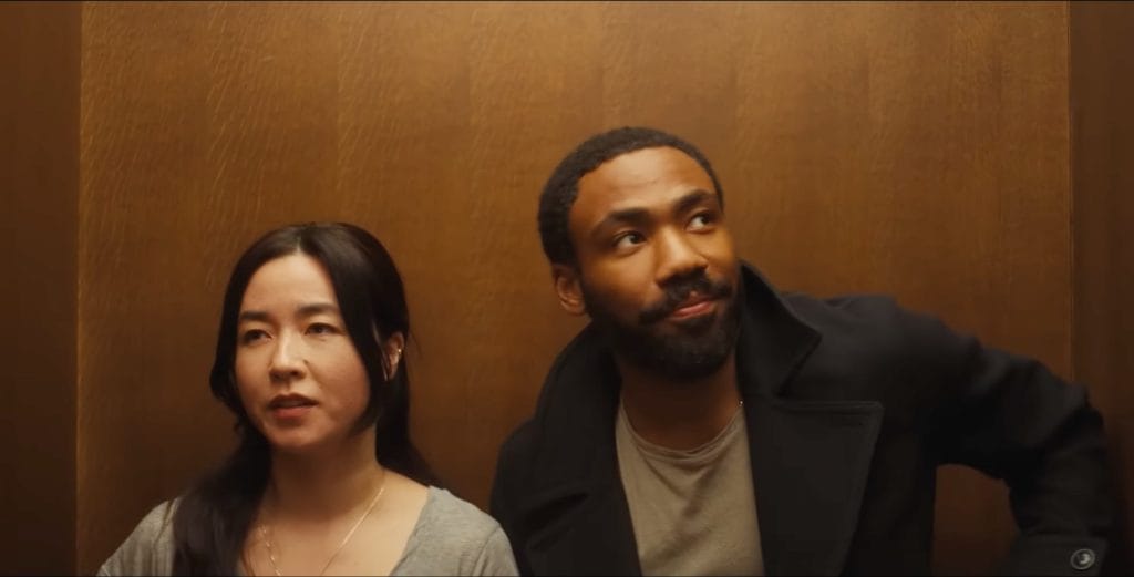 Glover and Esrkine standing next to each other in an elevator as they both look away from each other.
